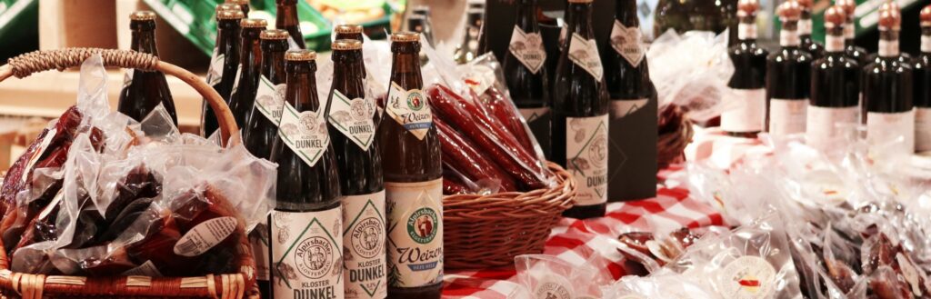 Biersorten und in Folie verpackte Wurstwaren auf rot-weiß dekorierter Tischdecke, teils in Korb, im Projektkontext von ‚Wilde Sau‘.