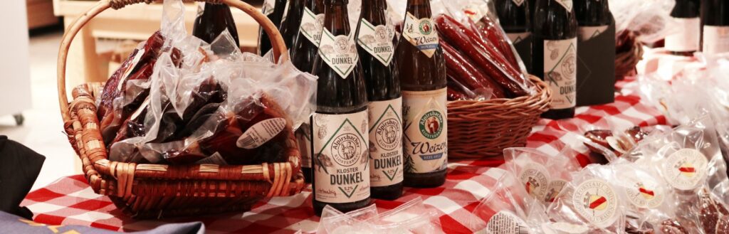 Markttisch mit Korb voller Wildschwein-Salami, Flaschen Bier mit 'Dunkle' Etikett, regionale Produkte auf rot-weiß kariertem Tuch.