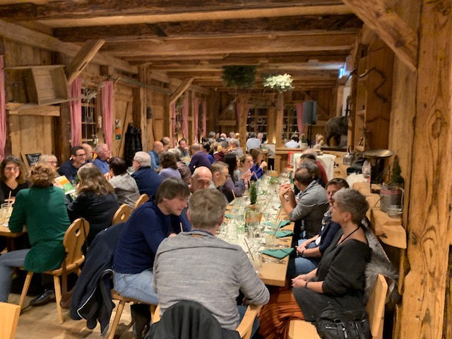 Menschen sitzen in rustikalem Saal an langen Tischen, trinken und essen. Holzbalken, Fenster mit roten Vorhängen sichtbar.