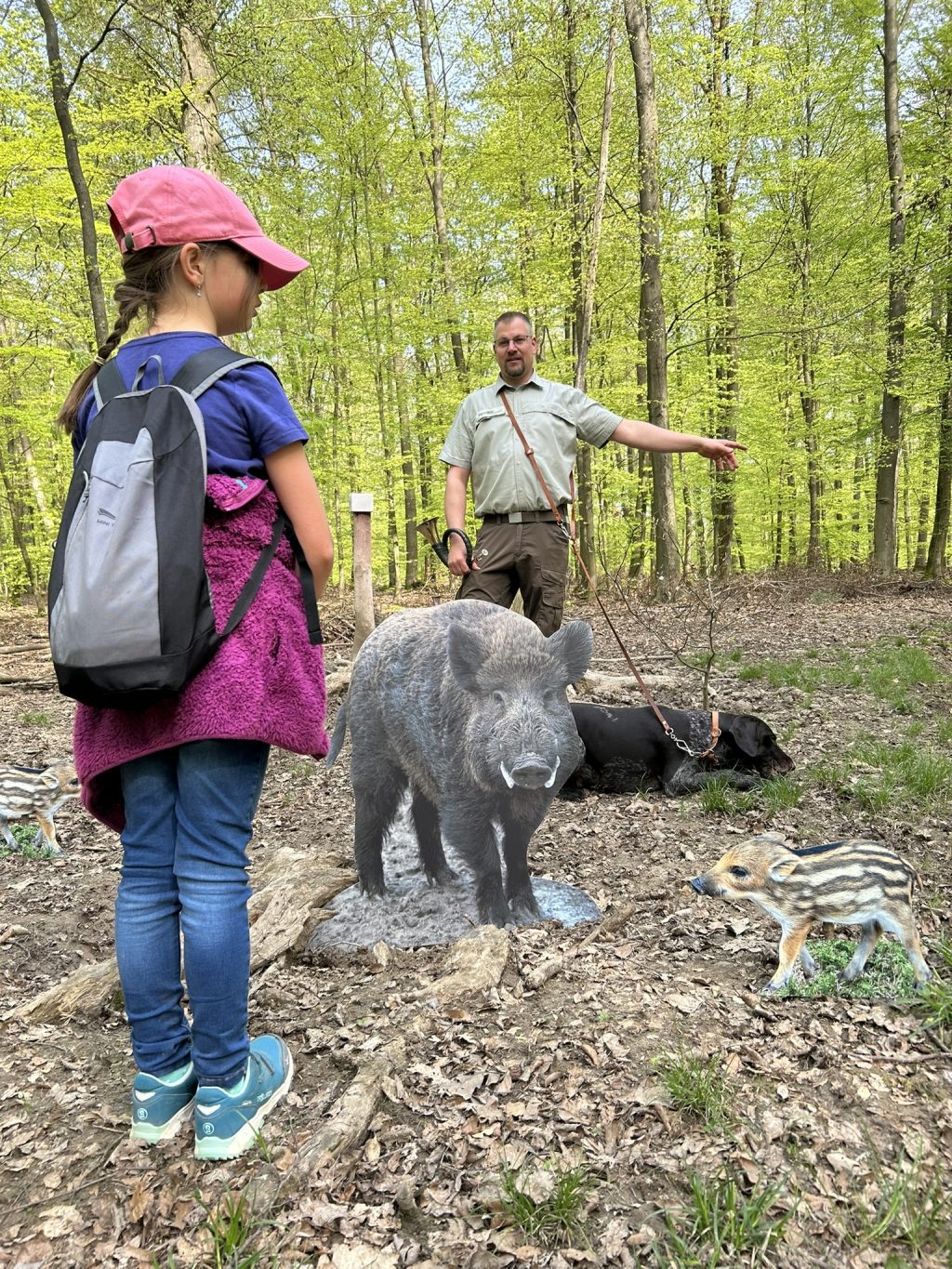 Kind mit Rucksack und Jäger bei einer Wildschweinskulptur im Wald stehend. Hund an Leine dabei.