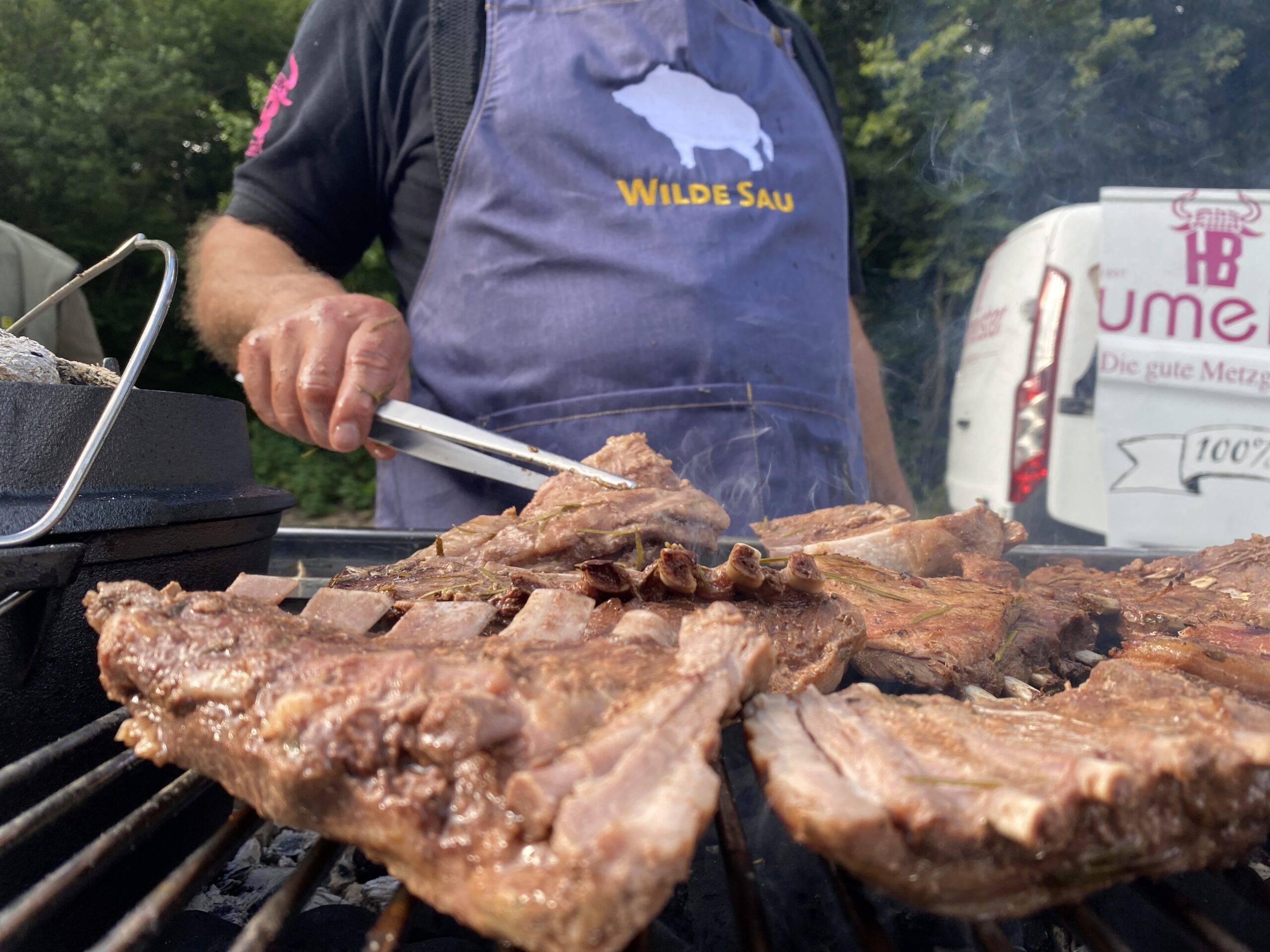 Gegrillte Wildschweinrippen, Mann mit Schürze und Grillzange, Lieferfahrzeug im Hintergrund.