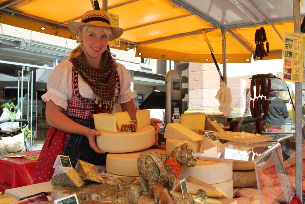 Frau in Tracht präsentiert Käse unter gelbem Marktstand, verschiedene Käsesorten und Salami am Stand.